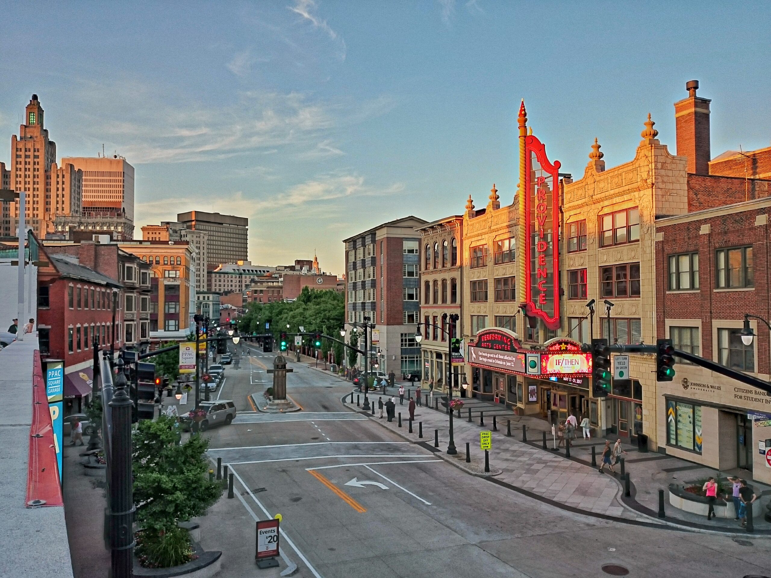 A city street at sunset with historic buildings, a large vertical theater sign that says "Theatre," and cars and pedestrians. Tall modern buildings are visible in the background under a clear sky.