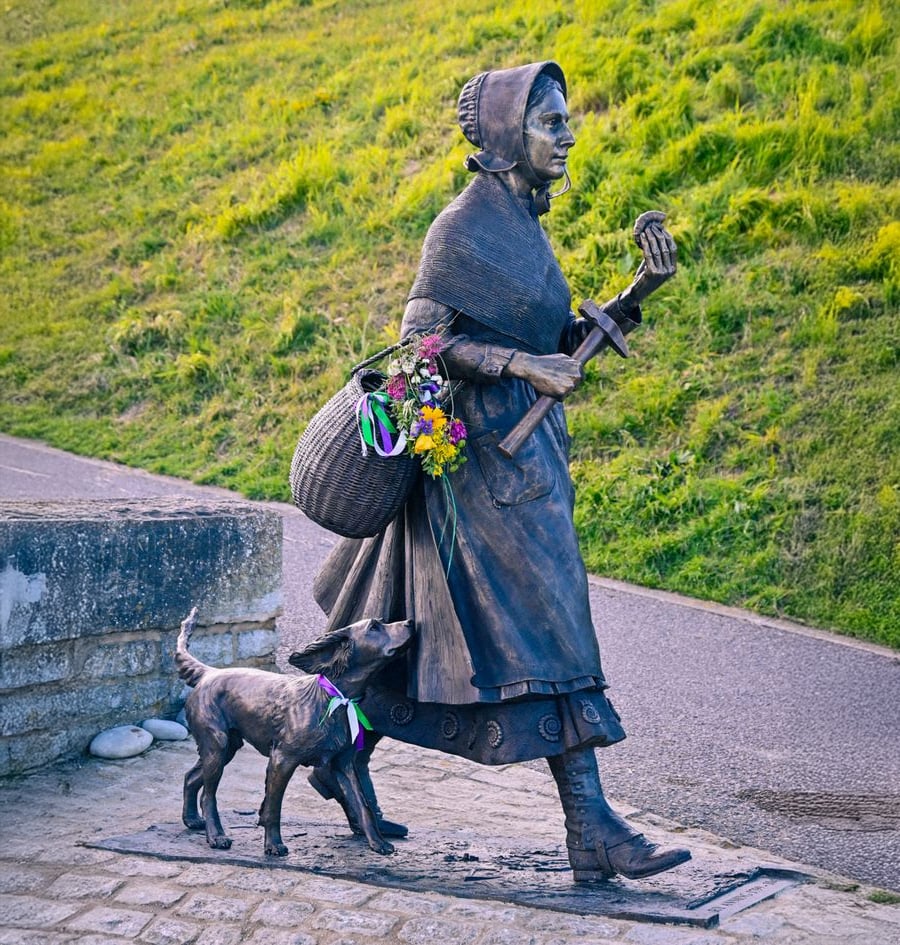 Bronze statue of a woman in historic clothing carrying a basket of flowers and a hammer, walking with a dog beside her on a pathway, set against a grassy hill.