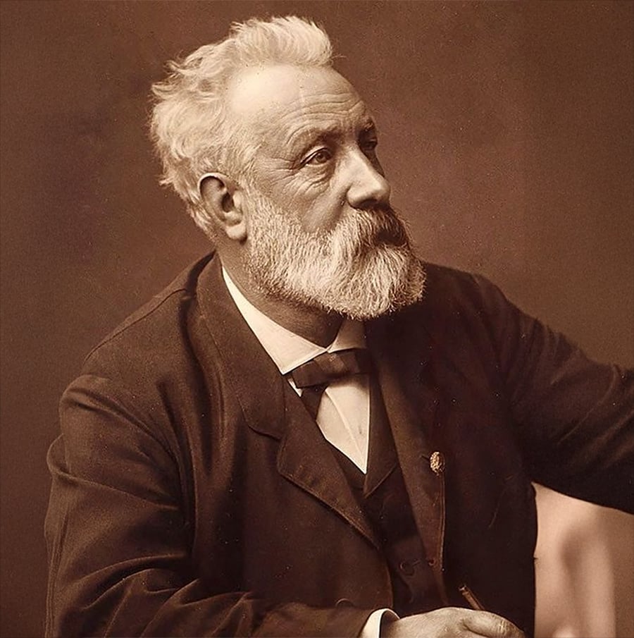 Older man with white hair and beard, wearing a dark suit and bow tie, sits and looks to the side against a plain background.