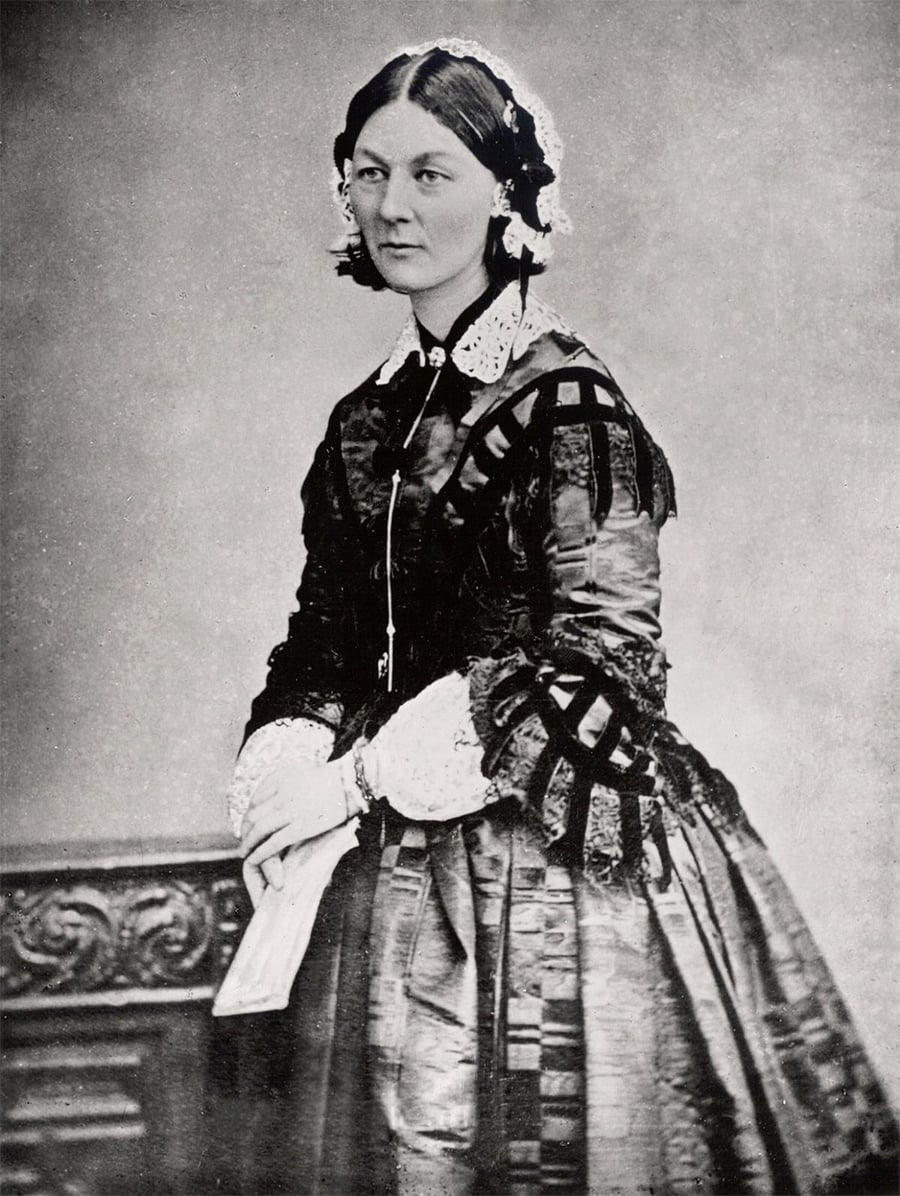 A black-and-white portrait of a woman in a Victorian dress, standing with her hands clasped and holding a paper, facing slightly left, with lace headpieces and a serious expression.