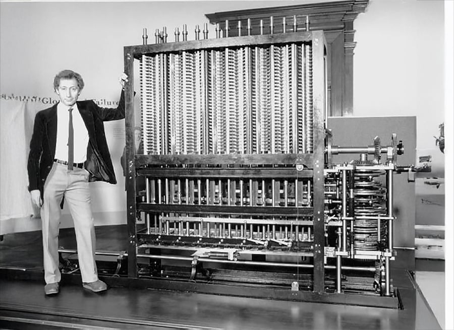 A man in a suit stands next to a large, complex mechanical machine with gears and columns, resembling an early computer or calculating device, in a room with minimal furnishings.