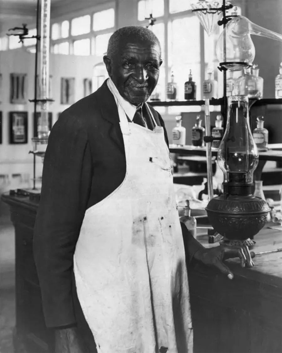 An older man in a suit jacket and apron stands in a laboratory, surrounded by glass bottles and scientific equipment, smiling slightly at the camera.