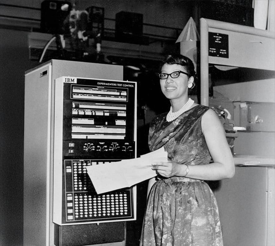 A woman in a dress and glasses stands beside an IBM computer, holding papers and smiling. The machine label reads “Experiments Test Control.” The setting appears to be a mid-20th century computing lab.