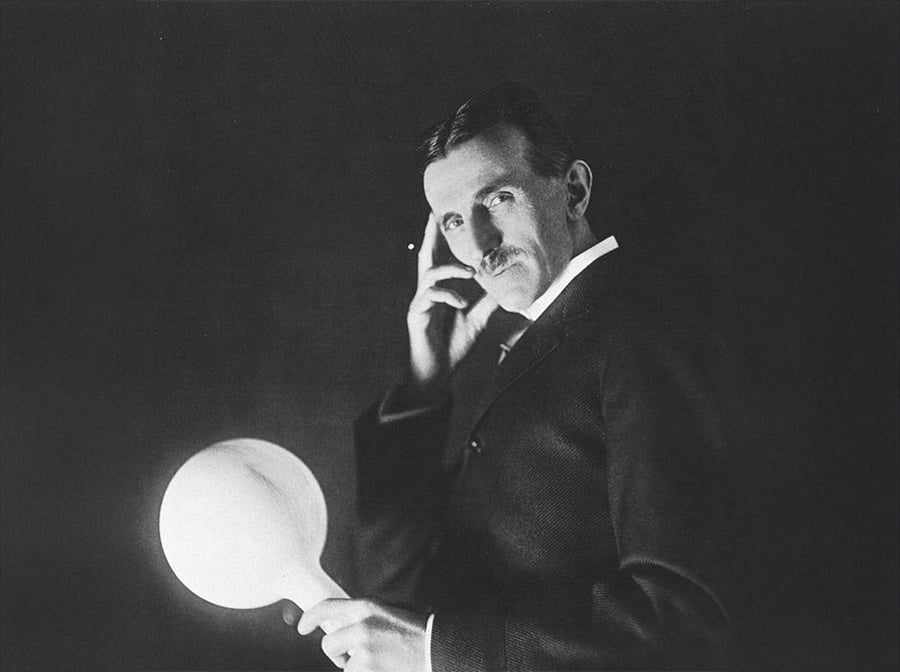A man with a mustache in a suit poses holding a glowing round object in one hand and touching his face with the other, set against a dark background.