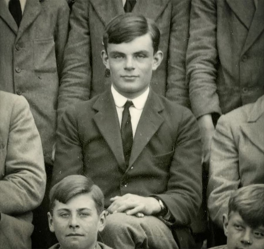 A young man in a dark suit sits with hands crossed in a group portrait, surrounded by others in similar attire. The photo is black and white and appears to be from the early 20th century.