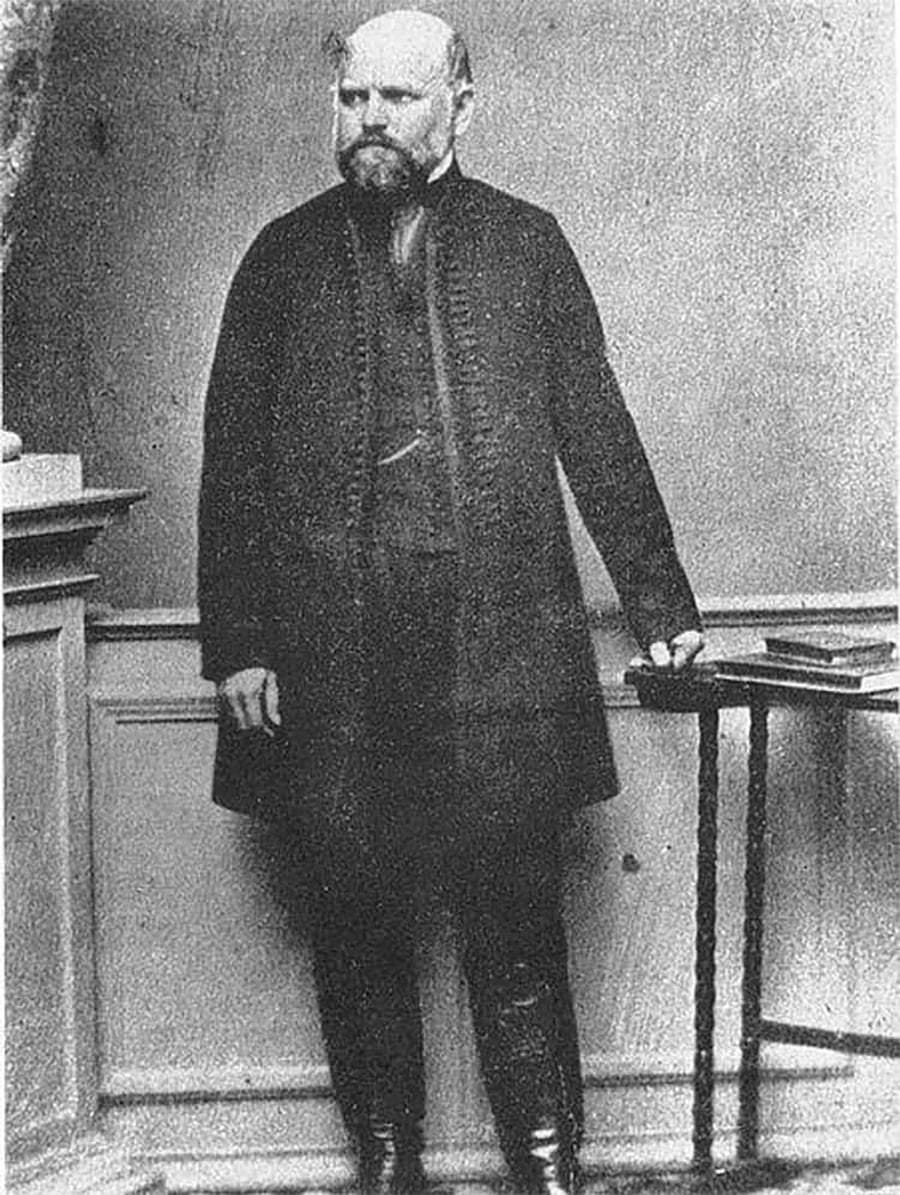 A bearded man stands indoors, wearing a long dark coat and boots, with one hand resting on a small table holding books. The photo is black and white and appears historical.