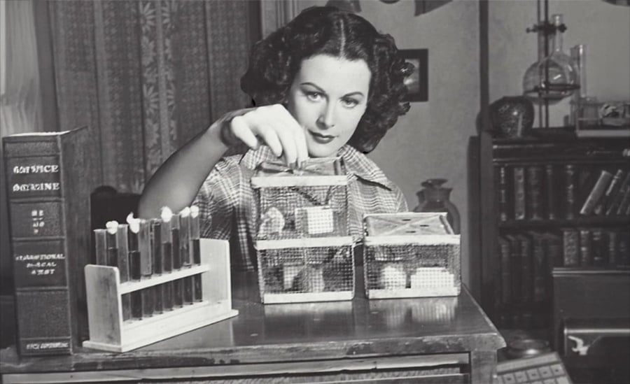 A woman in vintage clothing places an item in a mesh cage. Test tubes in a rack and a large science book labeled "Science Medicine" sit on a wooden table in front of her. Shelves and lab glassware are in the background.