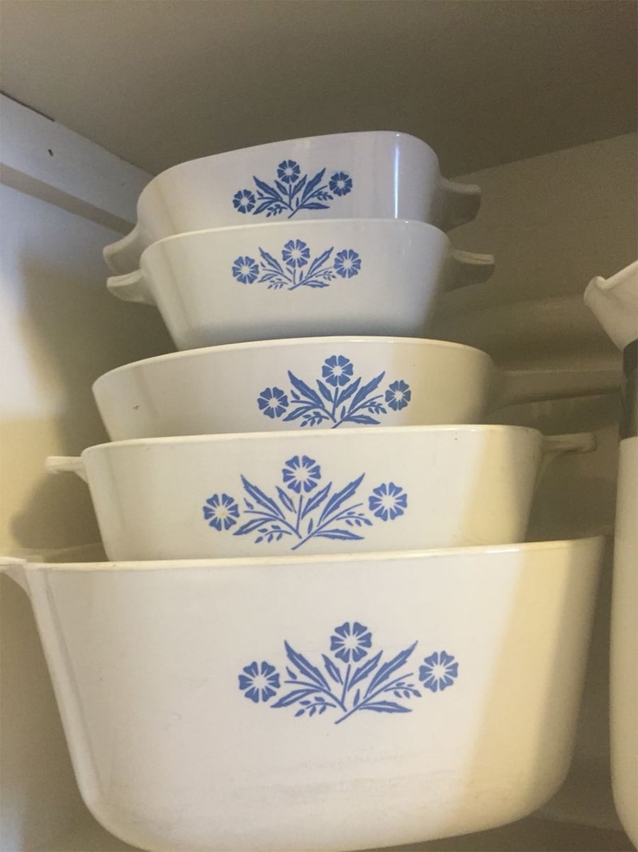 Four vintage white casserole dishes with blue floral patterns are nested together on a shelf inside a kitchen cabinet.