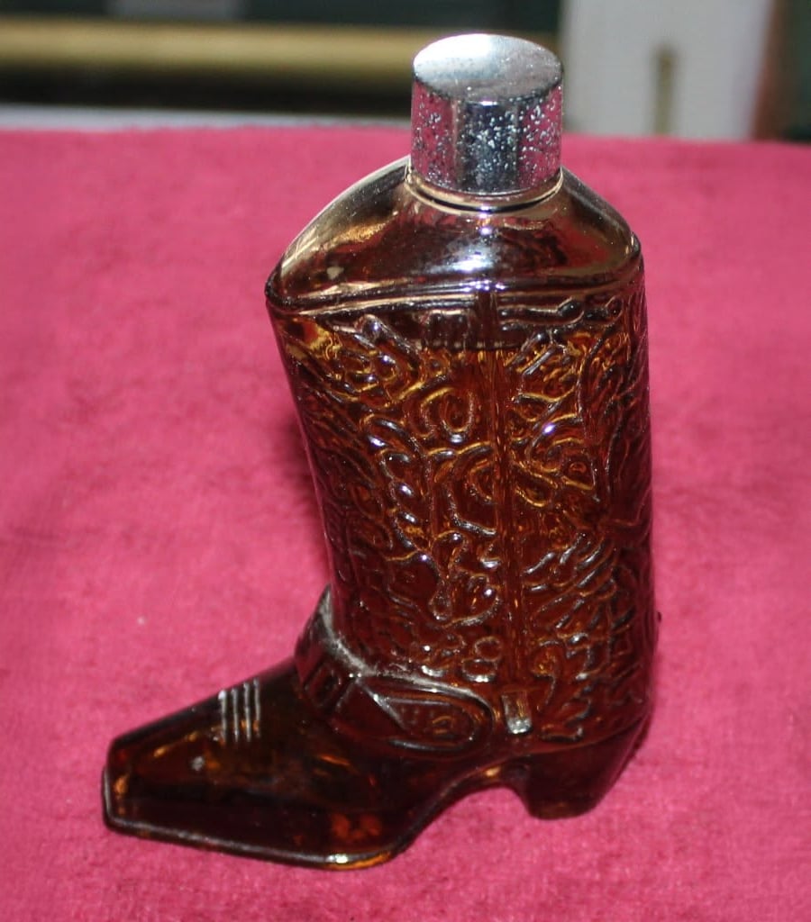 A brown glass bottle shaped like a cowboy boot with ornate detailing and a shiny silver cap, set on a red surface.