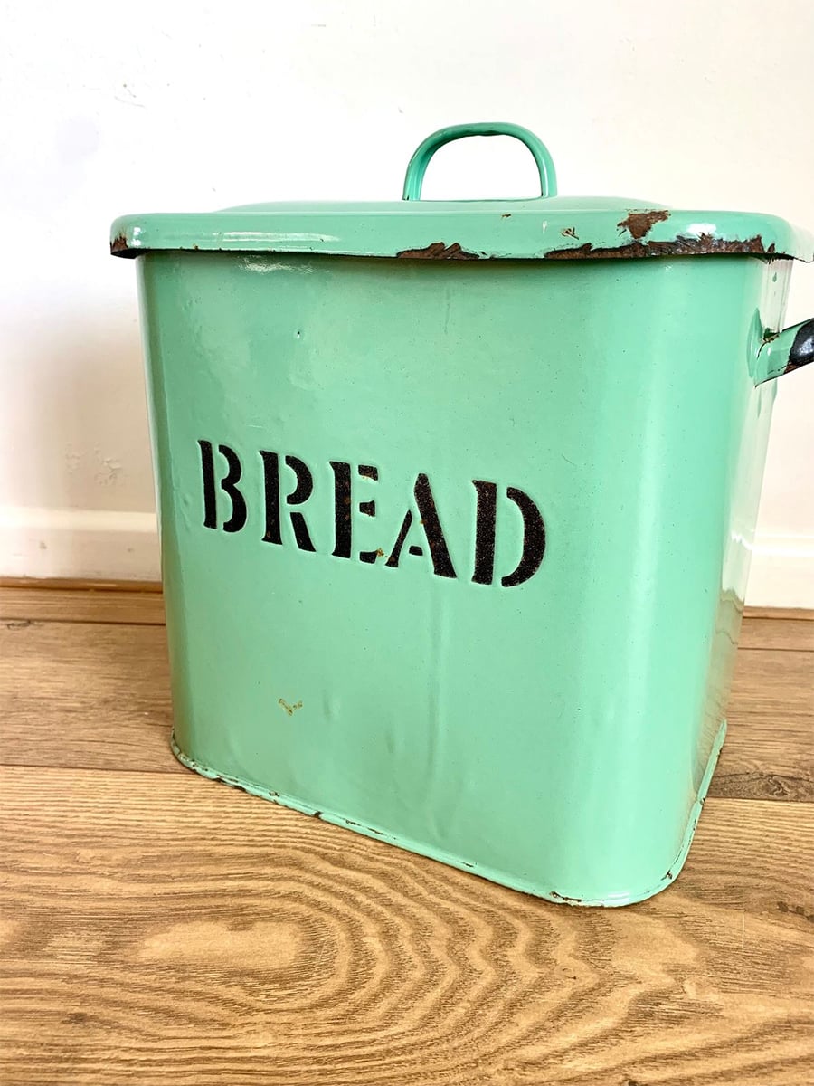 A vintage mint green metal bread box with a lid and the word "BREAD" stenciled in black sits on a wooden floor against a white wall. Some paint is chipped on the edges.
