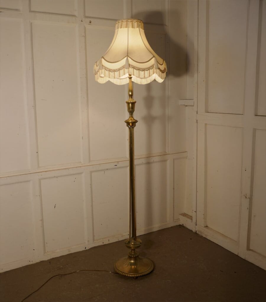 A vintage brass floor lamp with an ornate base and a cream-colored, scalloped fabric lampshade stands in the corner of a room with white paneled walls.