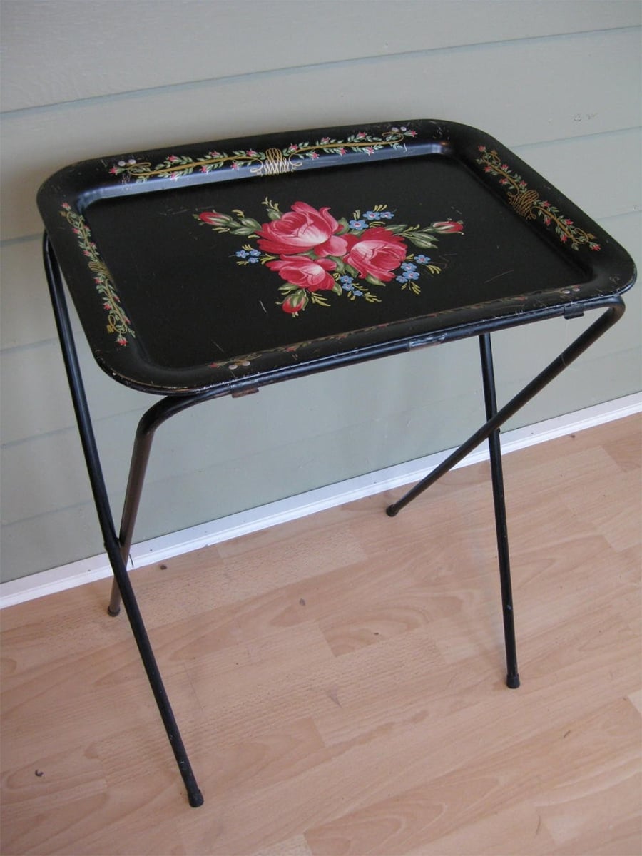 A vintage black metal TV tray with foldable legs, decorated with painted pink and red roses, sits on a light wood floor against a greenish-gray wall.