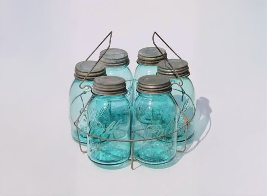 Five vintage blue Mason jars with metal lids are grouped together in a wire carrier, placed against a plain, light background.