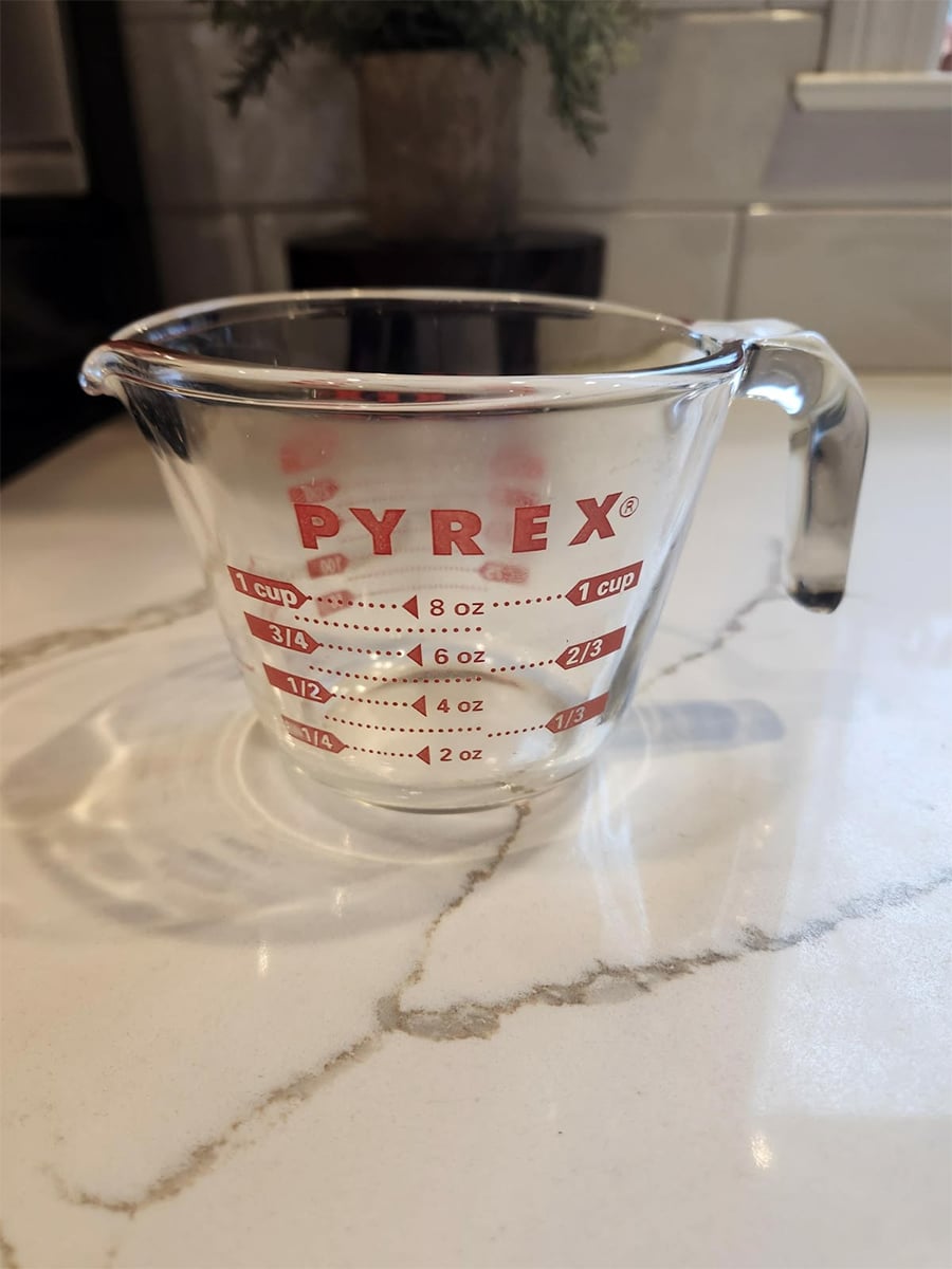 A clear Pyrex glass measuring cup sits on a marble countertop, showing red measurement markings for ounces and cups, with a handle and a spout for pouring.