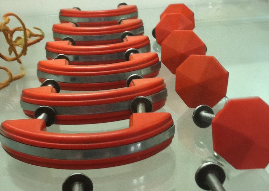 Four red octagonal dumbbells and four curved red hand grips with metal bands are displayed on a glass surface. A piece of dried plant is partially visible on the left side.