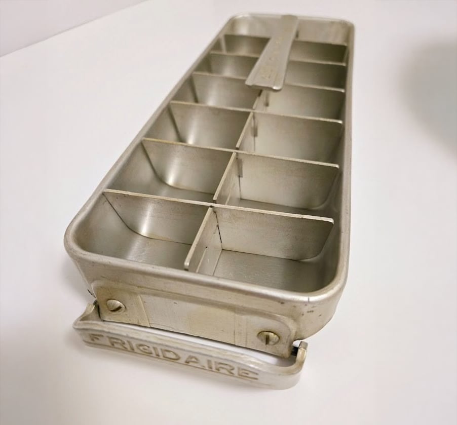 A vintage metal ice cube tray with a lever and dividers, labeled "Frigidaire" on the handle, sits on a white surface.