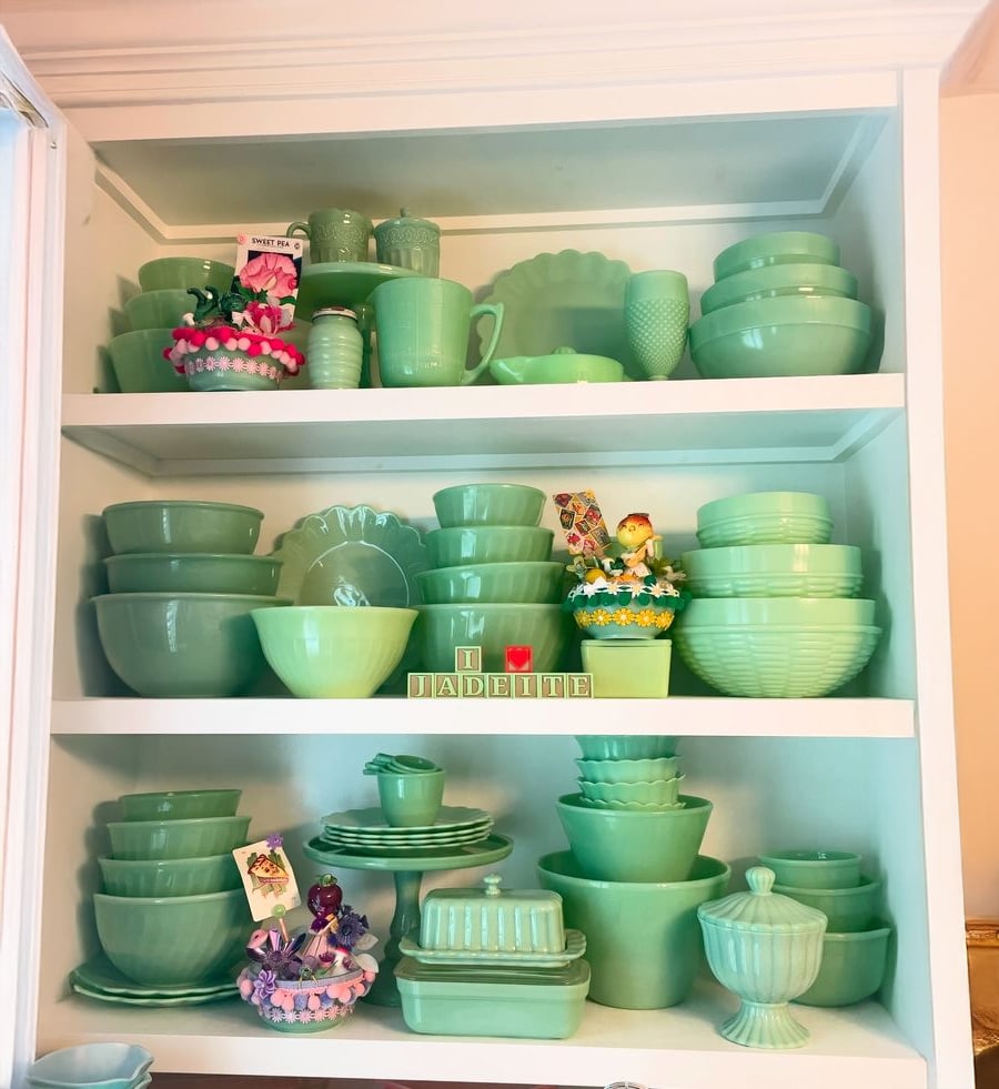 Three white shelves display a collection of vintage green jadeite dishes, bowls, cups, and decorative items. Two flower ornaments and a block sign reading “I ♥ JADEITE” are also visible.