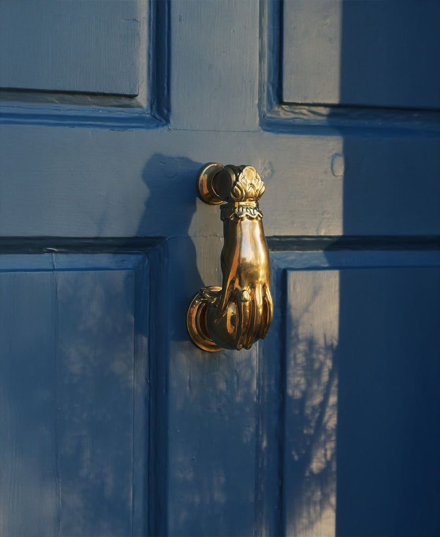A brass door knocker shaped like a hand is mounted on a blue wooden door. Shadows of branches or leaves are visible on the door’s surface near the knocker.