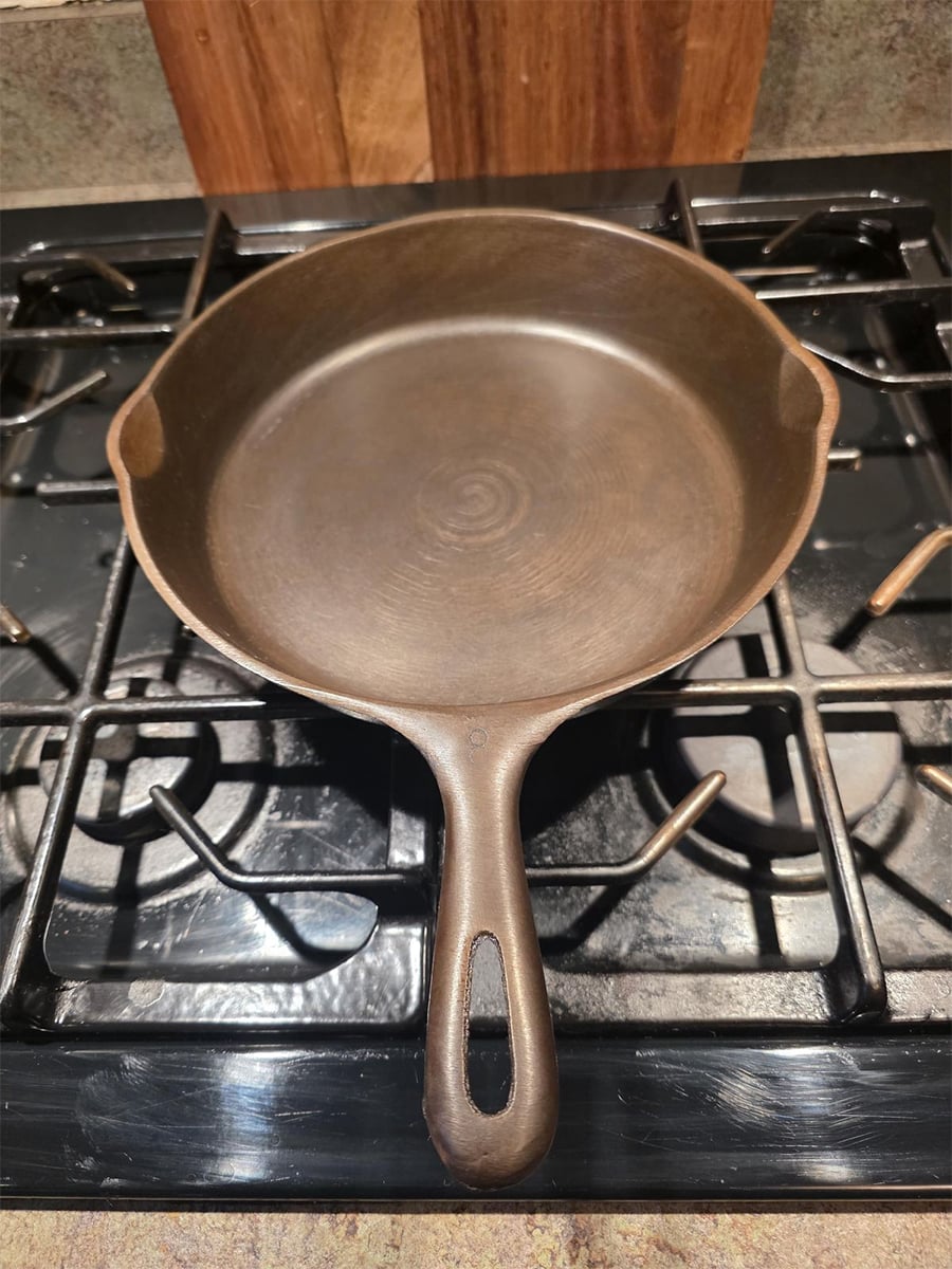 A round cast iron skillet with a handle is sitting on a black gas stove, surrounded by metal burners and grates. The skillet is empty and clean.