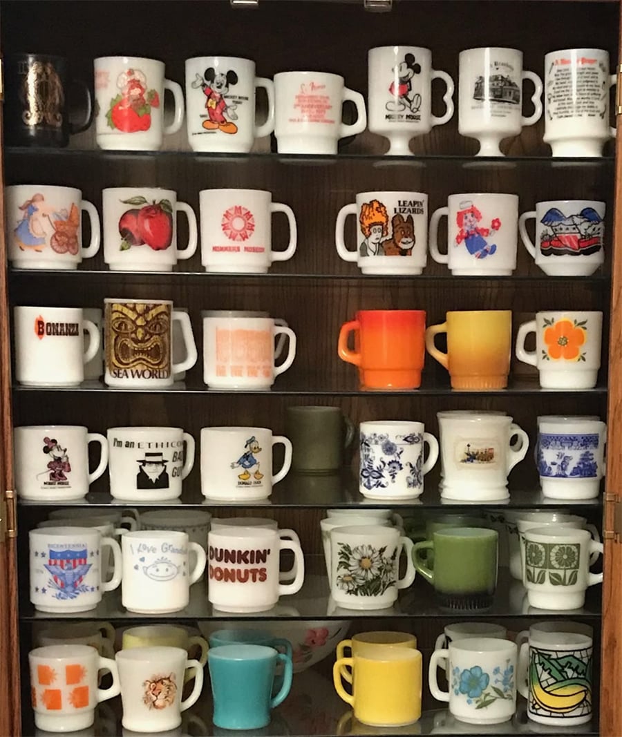 A cabinet displaying rows of vintage mugs, including cartoon characters, logos, floral patterns, restaurants, and colorful solid designs, all arranged neatly on shelves.