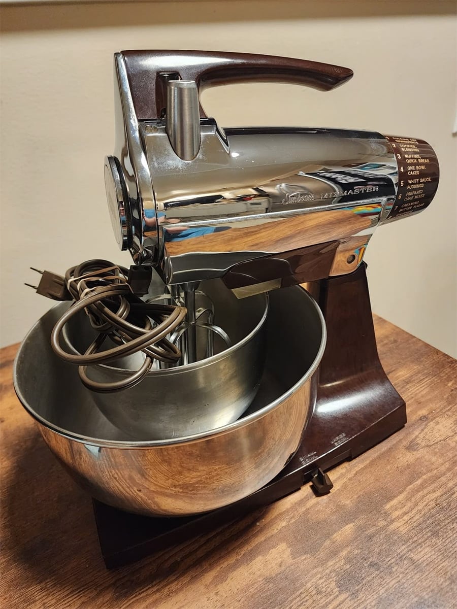 A vintage chrome stand mixer with a metal mixing bowl and attached electric cord sits on a wooden surface. The mixer features a dark brown base and handle, with multiple speed settings visible on the side.