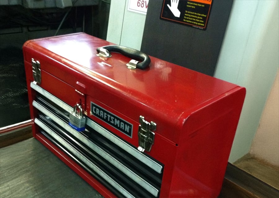 A red Craftsman toolbox with a padlock on the front sits on the floor next to a metal cabinet with a warning sign.