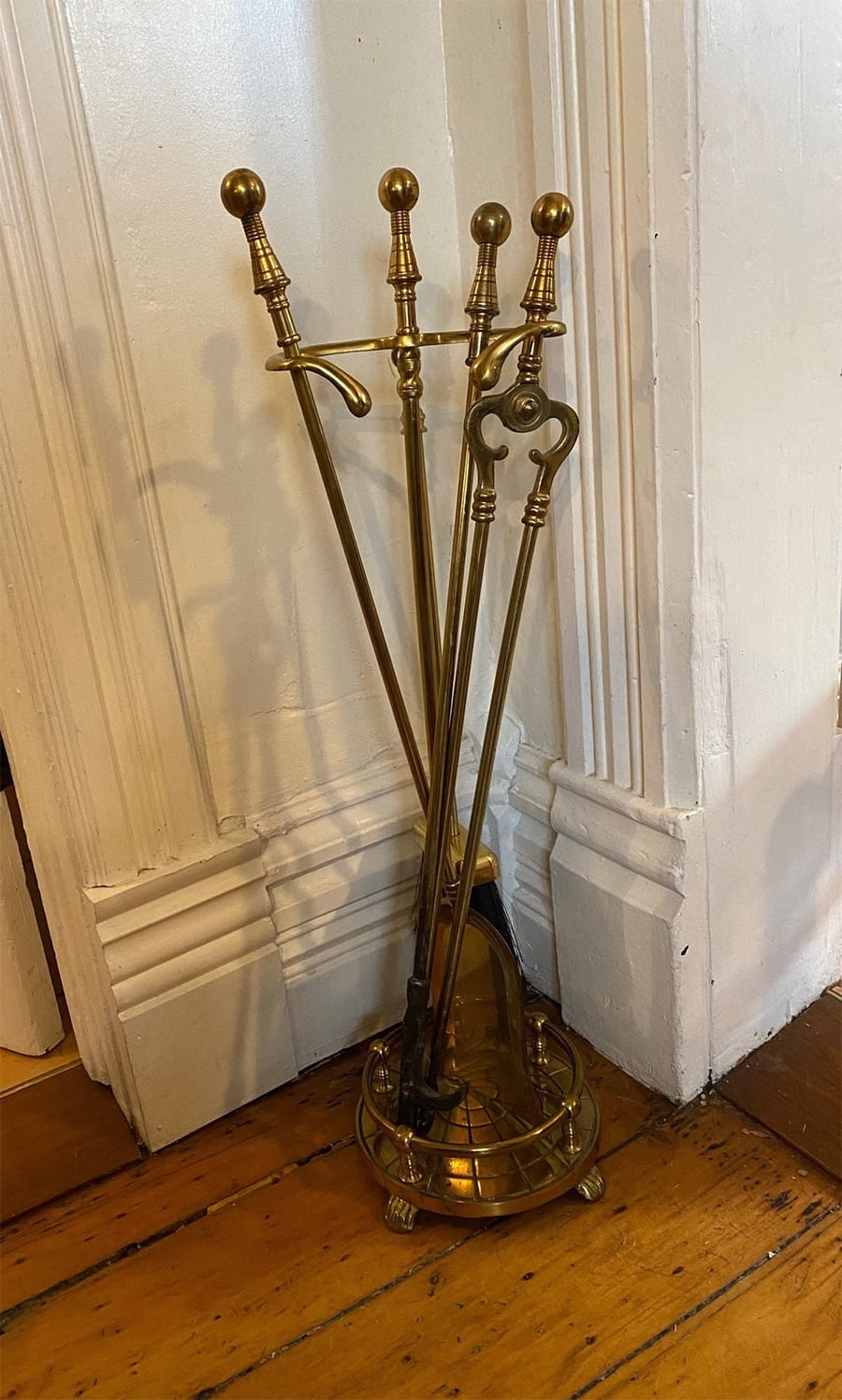A brass fireplace tool set with four tools—poker, tongs, brush, and shovel—stands on a matching brass holder against a white wall and wooden floor.