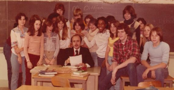 A vintage photo shows a teacher seated at a desk with books and papers, surrounded by smiling students in casual 1970s attire in a classroom with a chalkboard and holiday decorations.