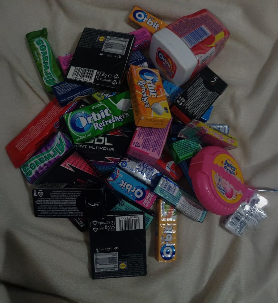 A pile of assorted chewing gum packs and containers, including Orbit, 5 Gum, Mentos, Bubblicious, and Juicy Fruit, displayed on a light-colored blanket.