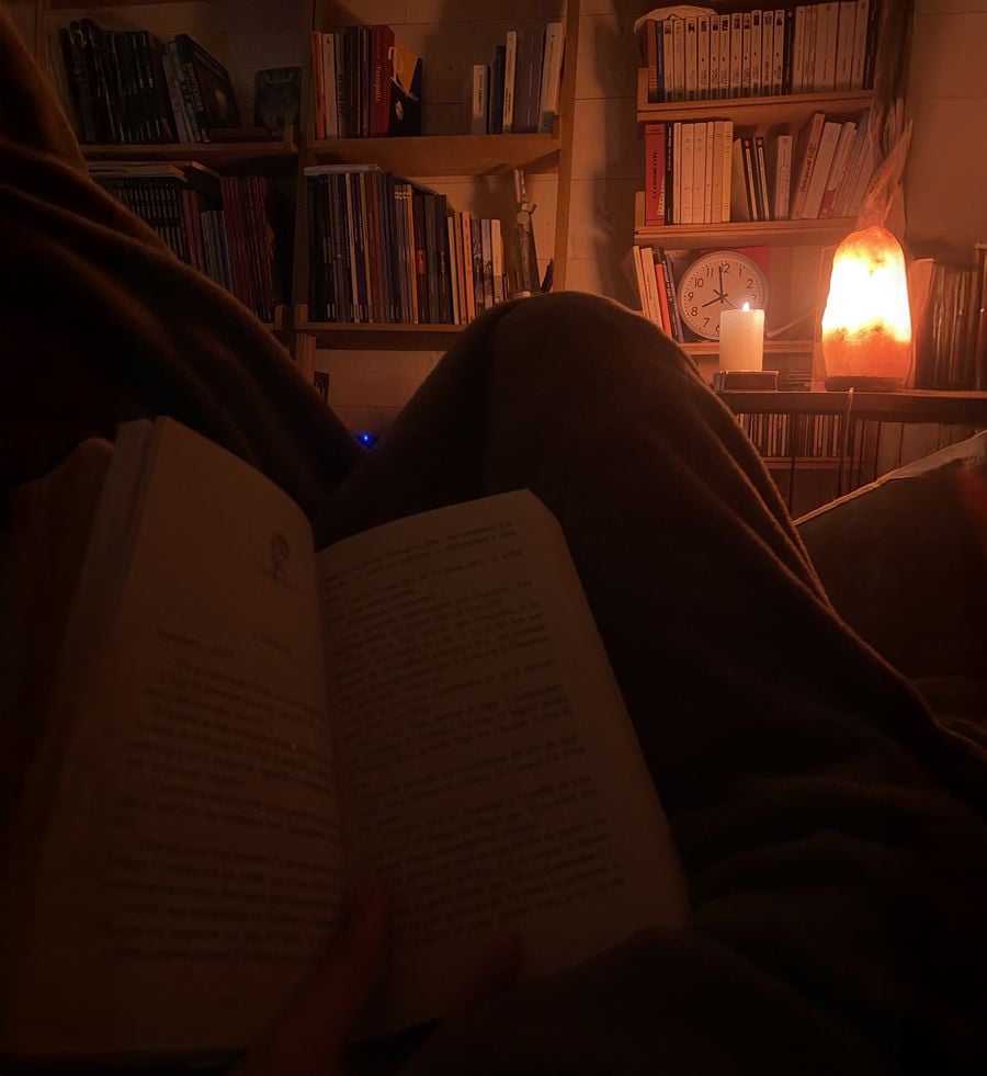 A person reads a book in a dimly lit room with bookshelves, a salt lamp, a candle, and a clock in the background, creating a cozy and warm atmosphere.
