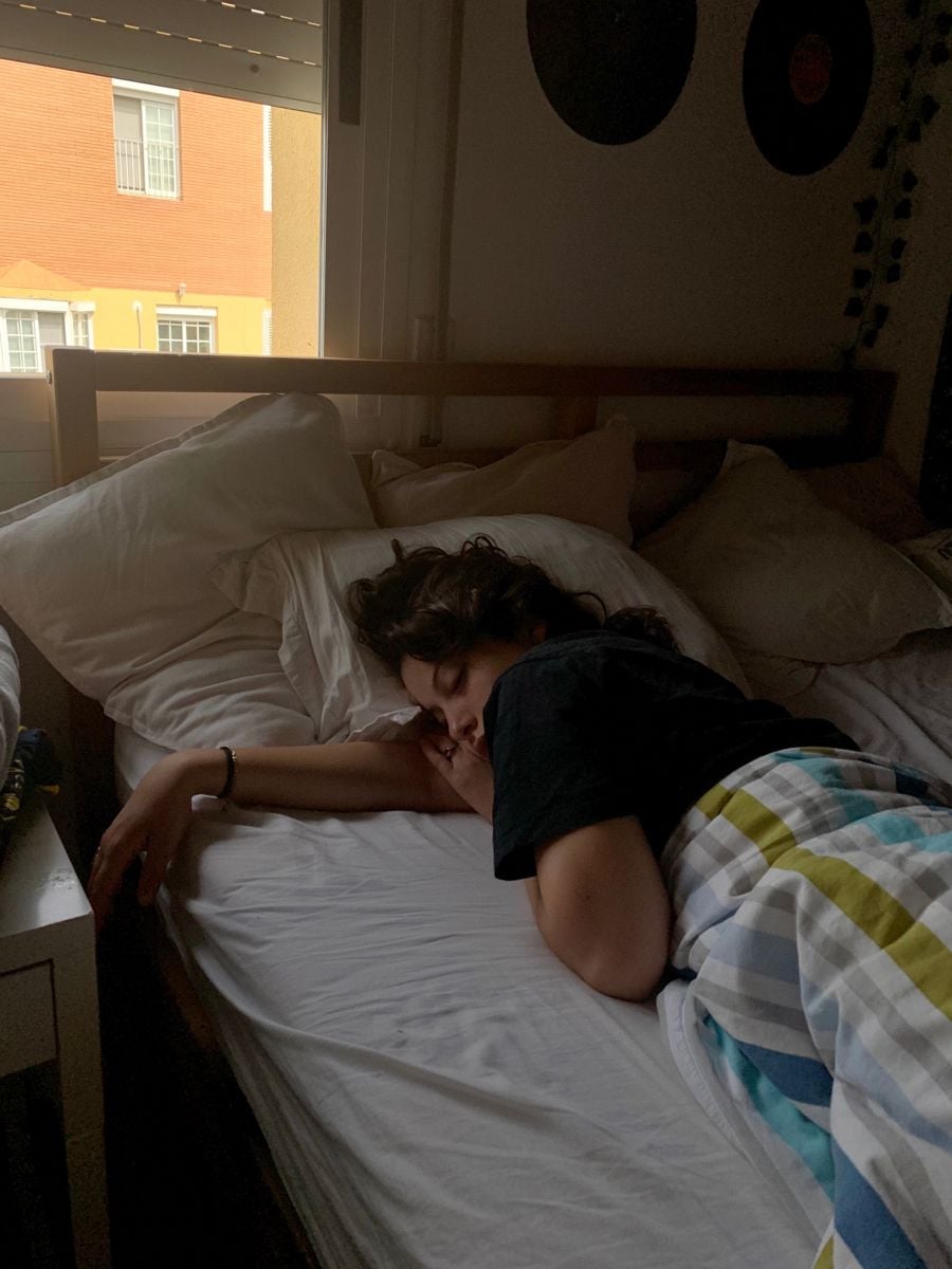 A person with dark hair sleeps on a bed by a window, covered with a striped blanket. Light from outside softly illuminates the room, and vinyl records hang on the dark wall in the background.