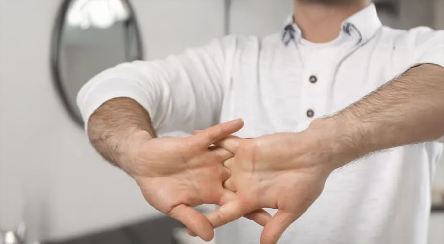 A person in a white shirt is stretching their fingers and knuckles with both hands clasped together, standing indoors near a round mirror.