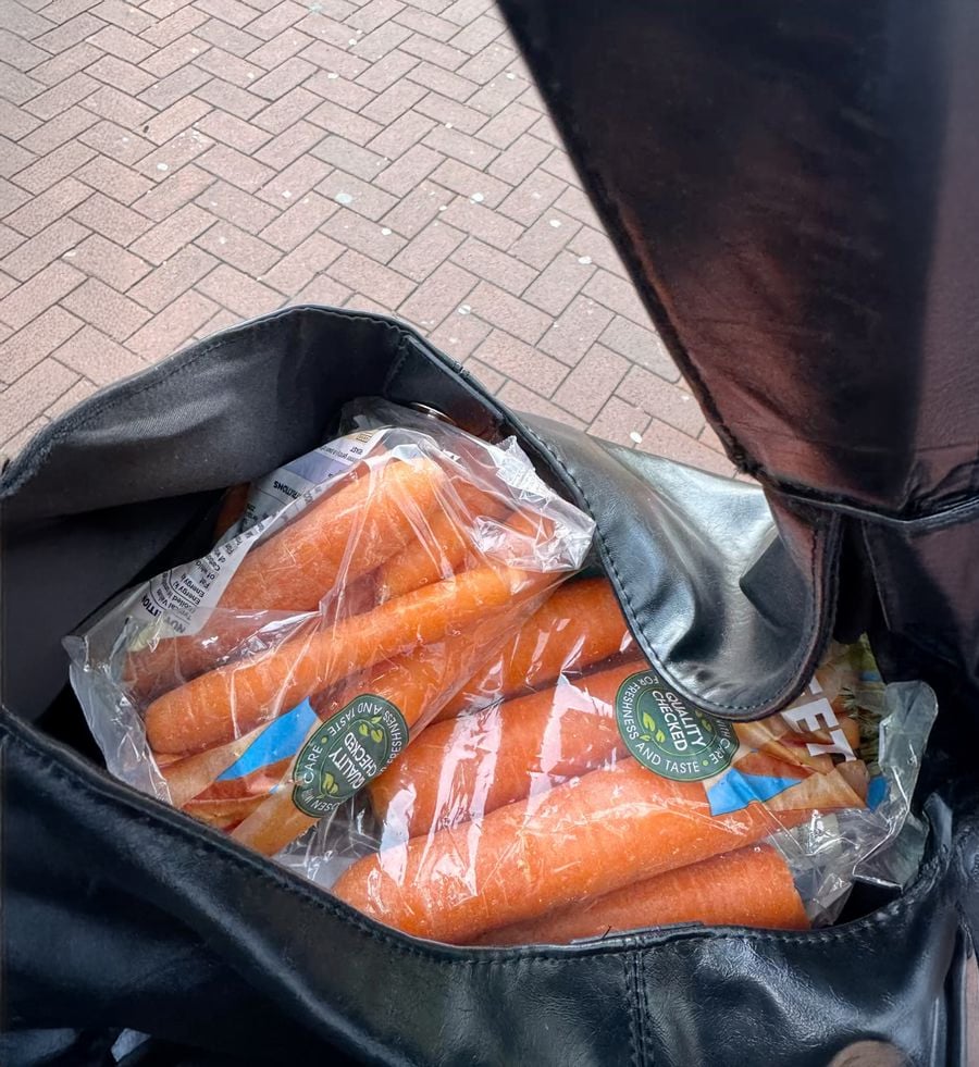 A black leather bag is open, revealing two plastic bags filled with whole carrots inside. The setting appears to be outdoors on a brick pavement.