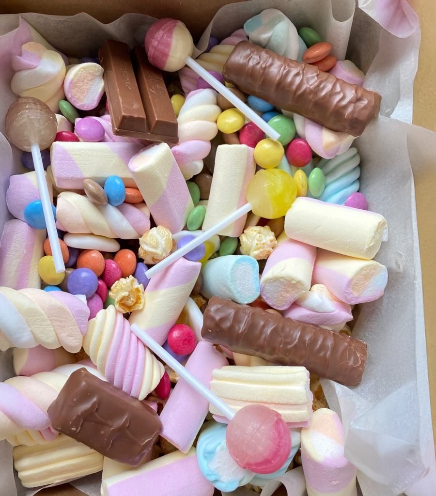 Assorted colorful candies, marshmallows, chocolate bars, lollipops, and pastel sweets in a paper-lined box.