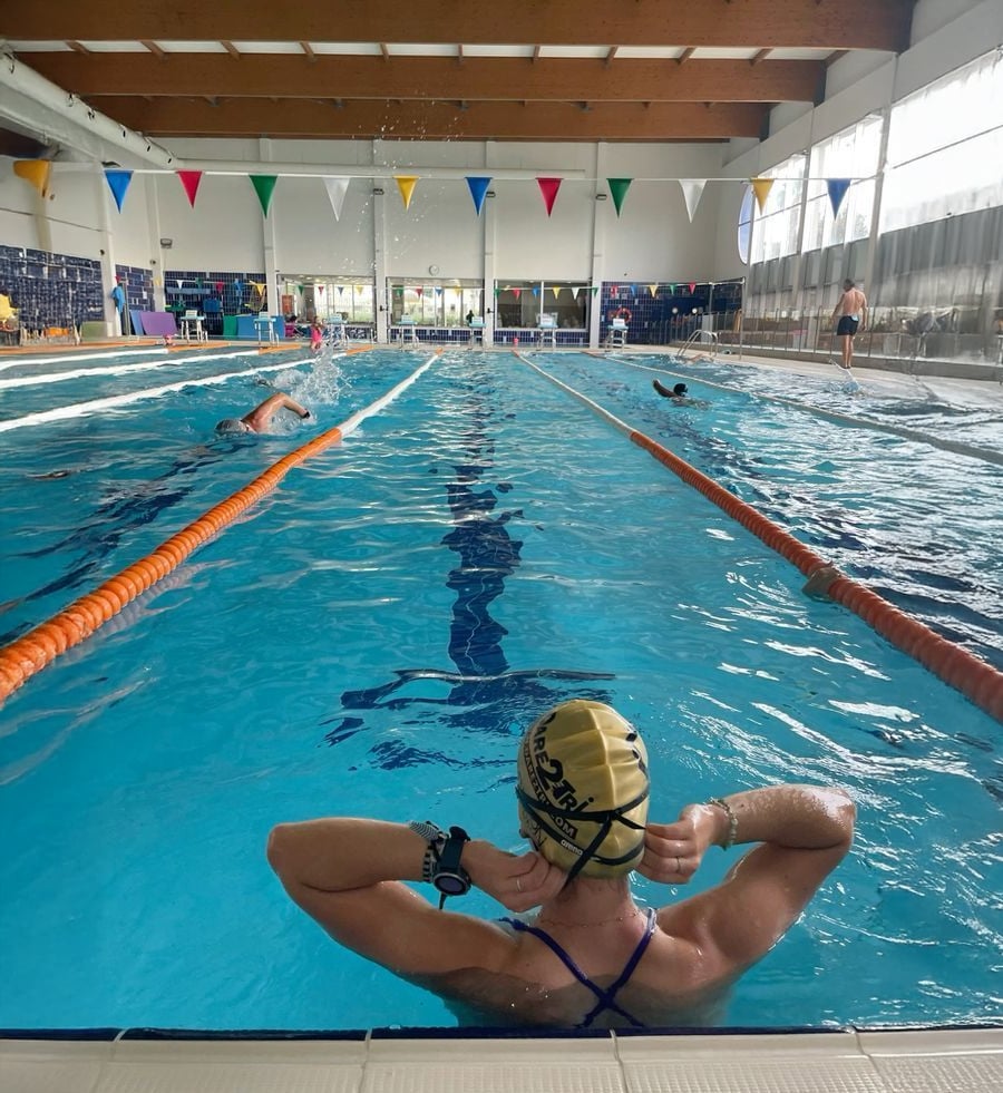 A swimmer wearing a cap and goggles sits at the edge of an indoor pool, preparing to enter. Other swimmers are swimming laps in separate lanes. Colorful flags hang overhead.