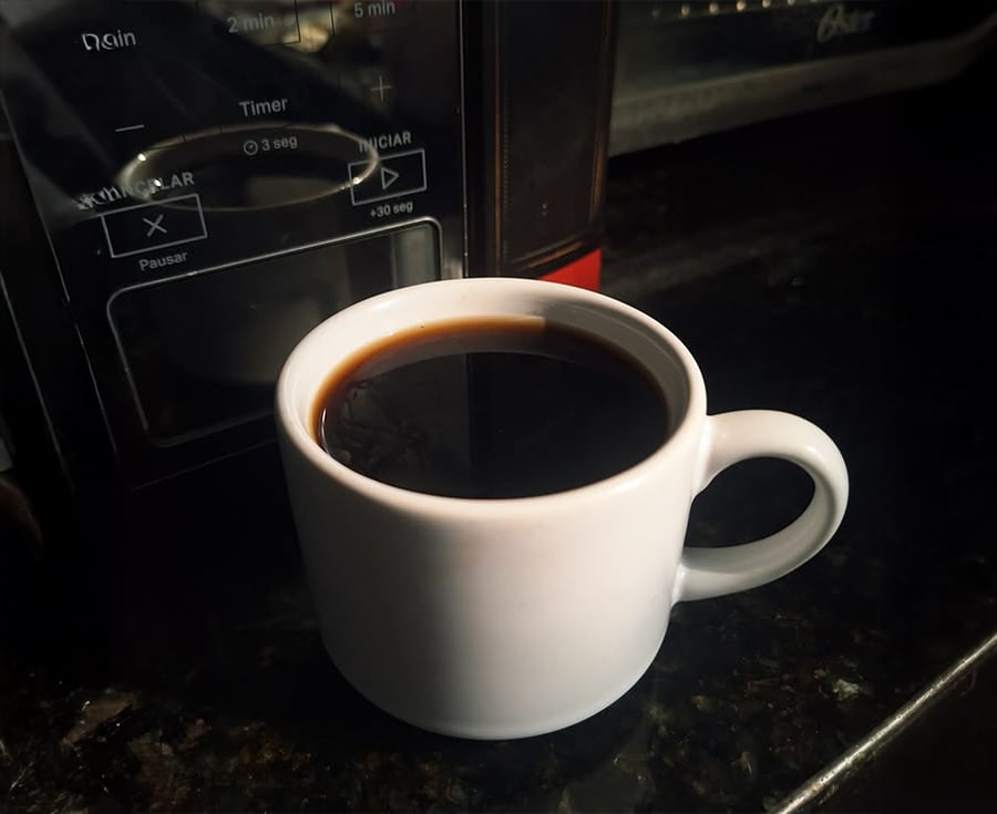 A white ceramic mug filled with black coffee sits on a dark countertop in front of a microwave oven.