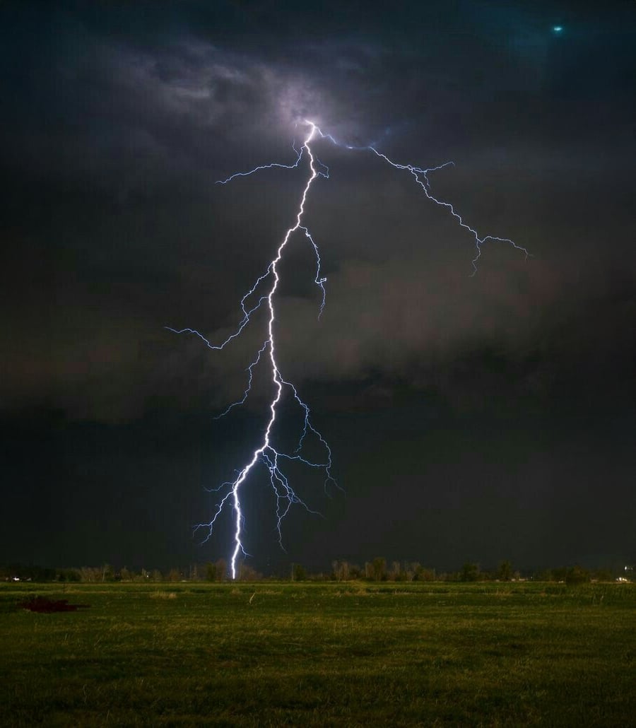 A bolt of lightning strikes from dark storm clouds onto a grassy field at night, illuminating the sky with bright, branching tendrils.