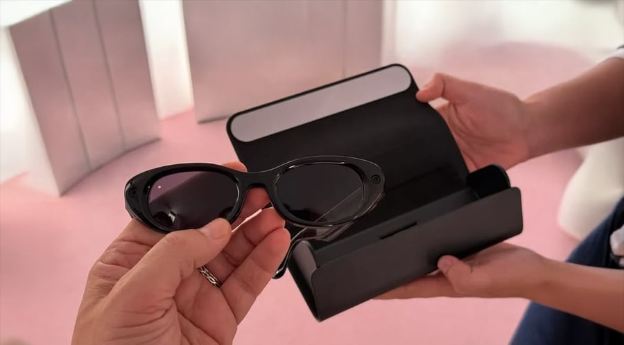 A person holds smart glasses with dark lenses, while another person opens a black glasses case against a pink background.
