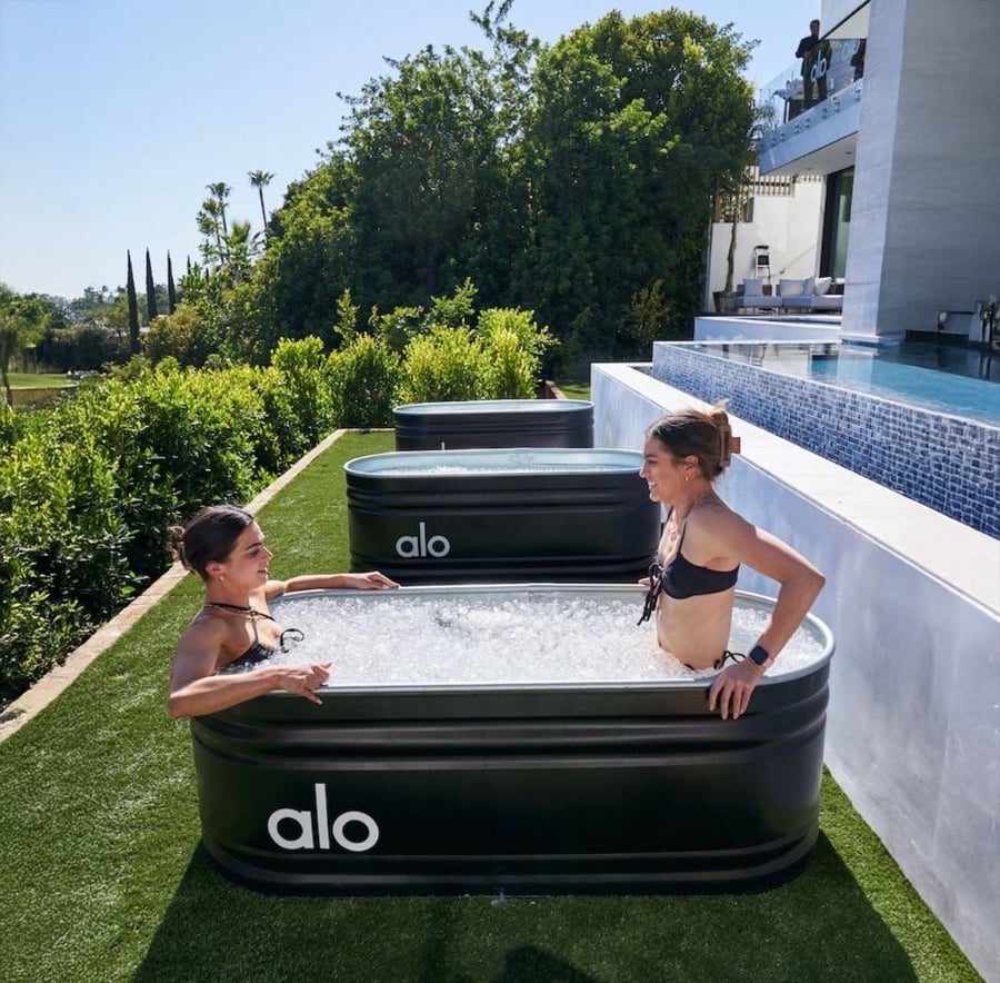 Two women in swimsuits sit in separate black alo ice baths on grass next to a pool, with bushes, trees, and a modern white building in the background on a sunny day.