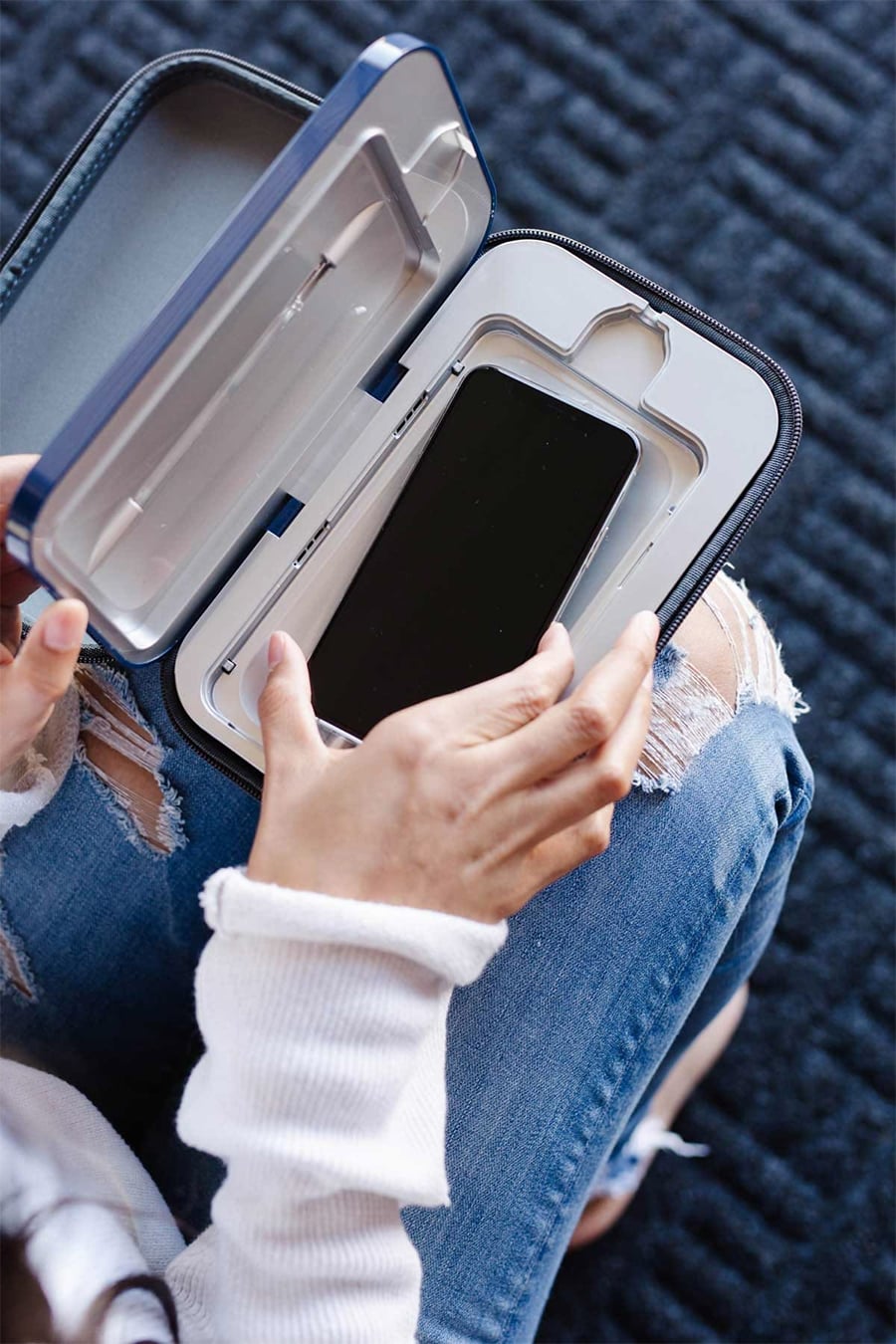 Person placing a smartphone into a UV sanitizer case while sitting on a chair, wearing ripped blue jeans and a white sweater.