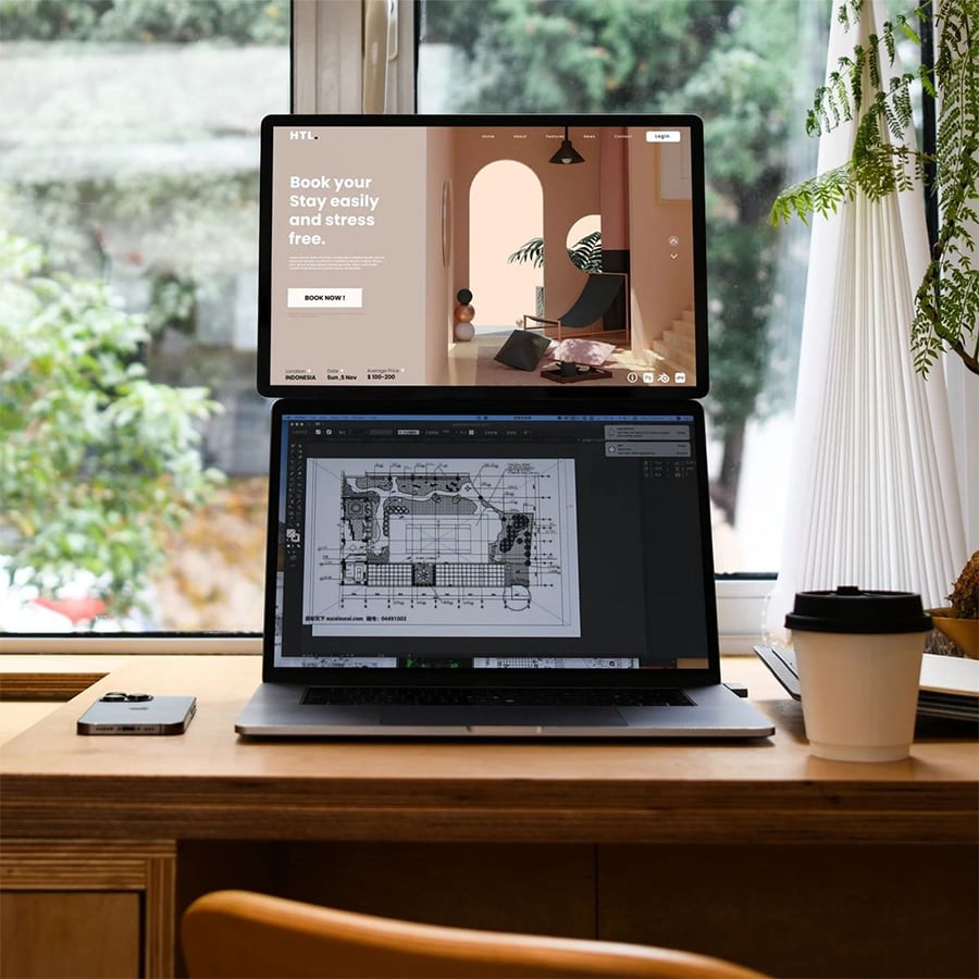 A laptop with two stacked screens displays a website and architectural plans on a wooden desk by a window, with a coffee cup, phone, and potted plant nearby.