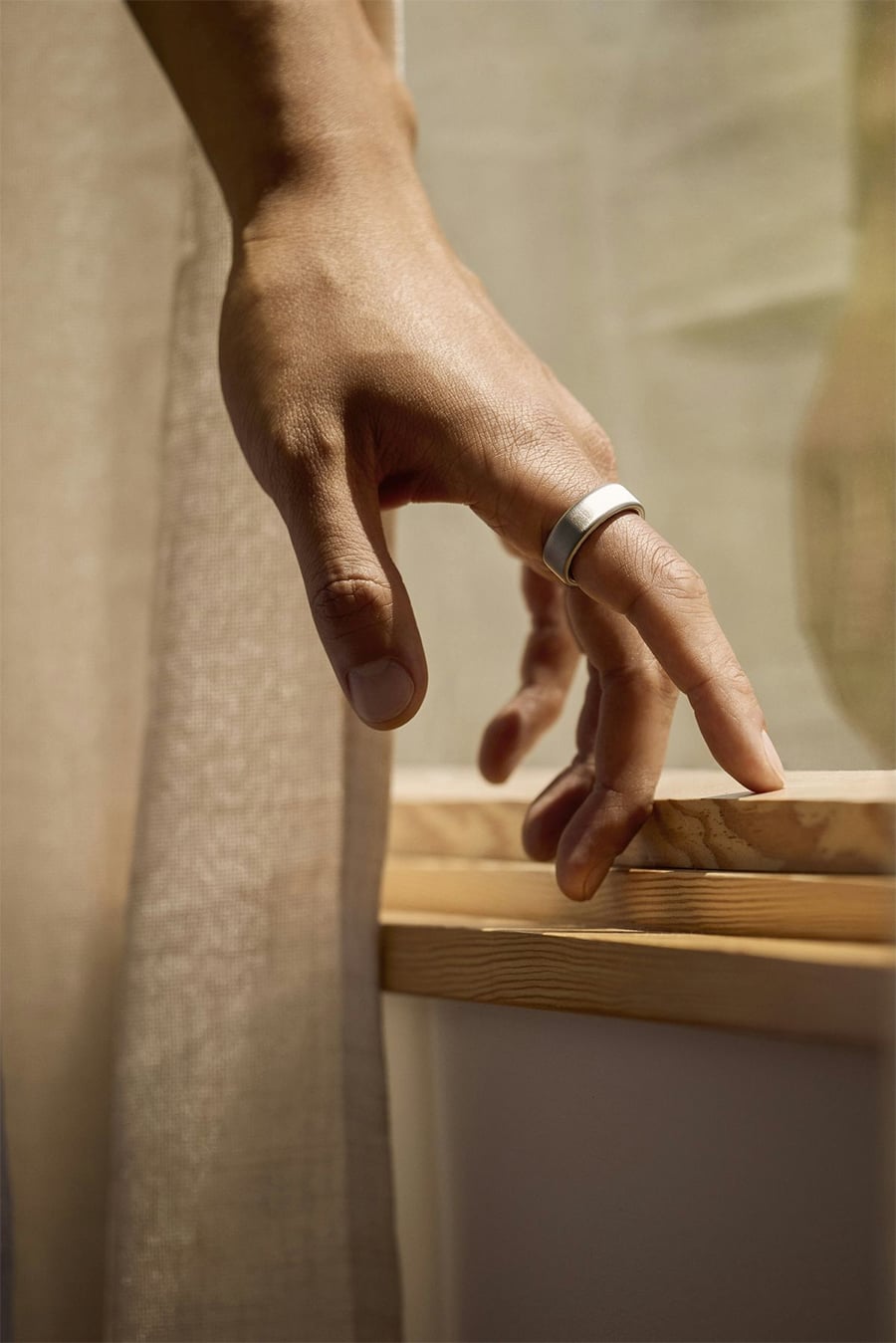 A hand with a silver ring rests gently on a windowsill, next to a beige curtain, with soft natural light streaming in.