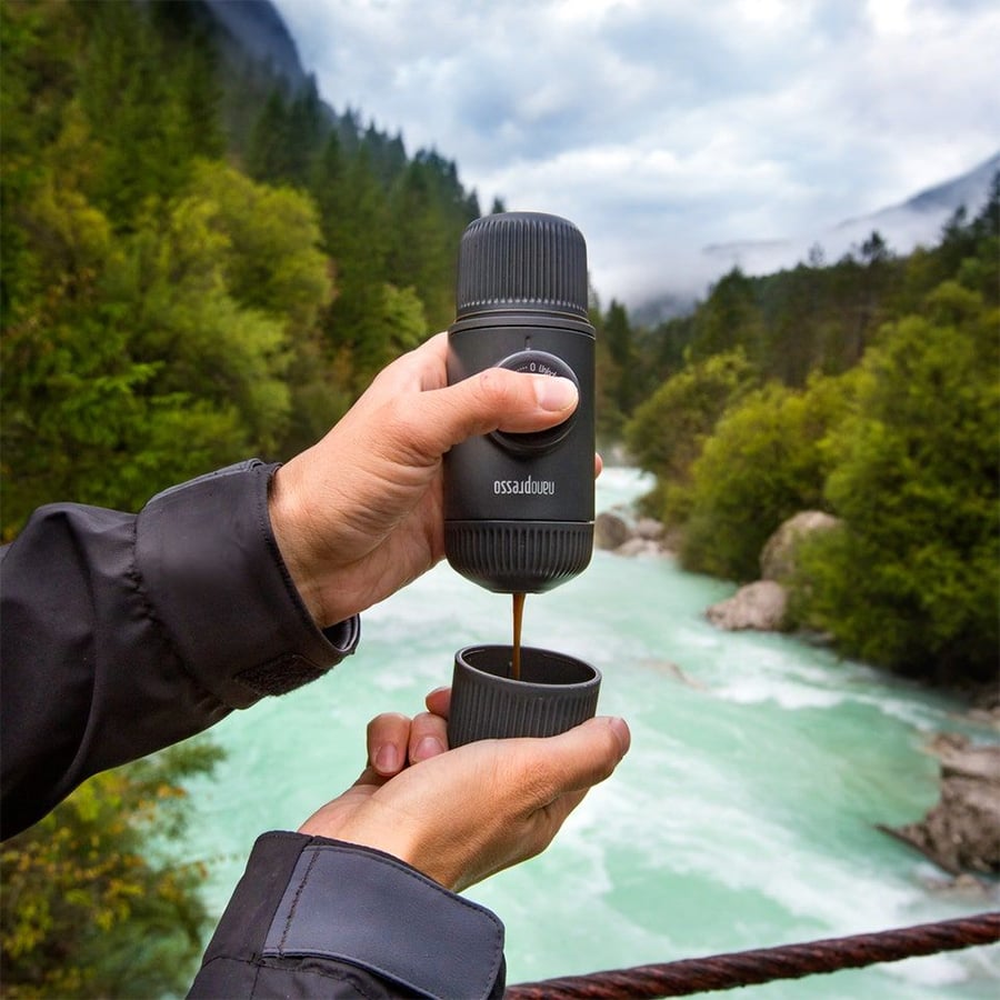 A person uses a portable coffee maker to pour coffee into a small cup, with a scenic river, trees, and mountains in the background.
