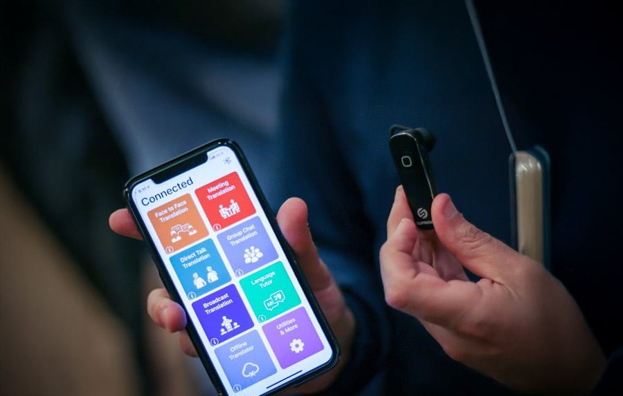 A person holds a smartphone displaying a colorful app and a small black electronic device with a button, wearing a lanyard with card in background.