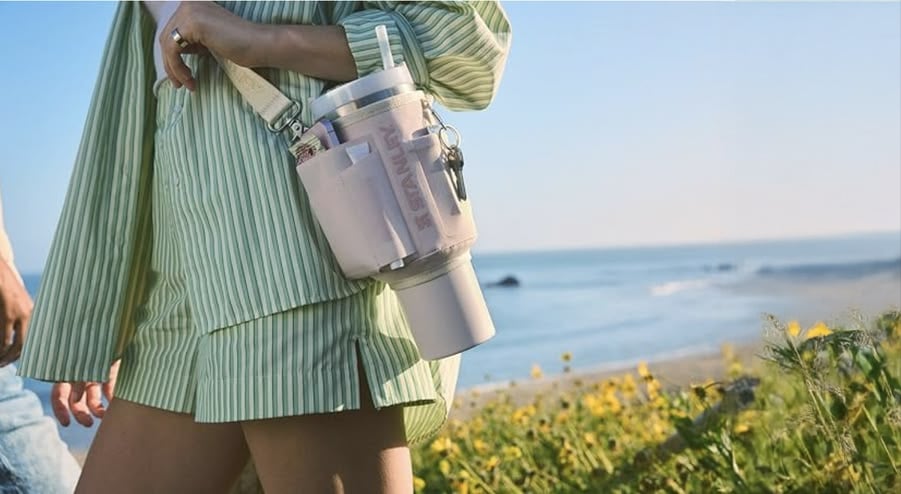 Person in a green striped shirt and shorts stands by the beach, carrying a large insulated tumbler with keys and a crossbody strap. Wildflowers and ocean waves are visible in the background.