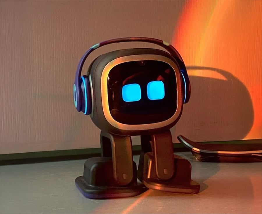 A small robot with a square face, glowing blue eyes, and wearing blue headphones stands on a table, lit by warm orange and yellow light.
