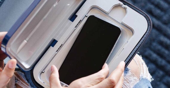A person places a smartphone inside a portable sanitizing device, possibly for UV cleaning. The device is white and open, and the person is wearing ripped jeans.