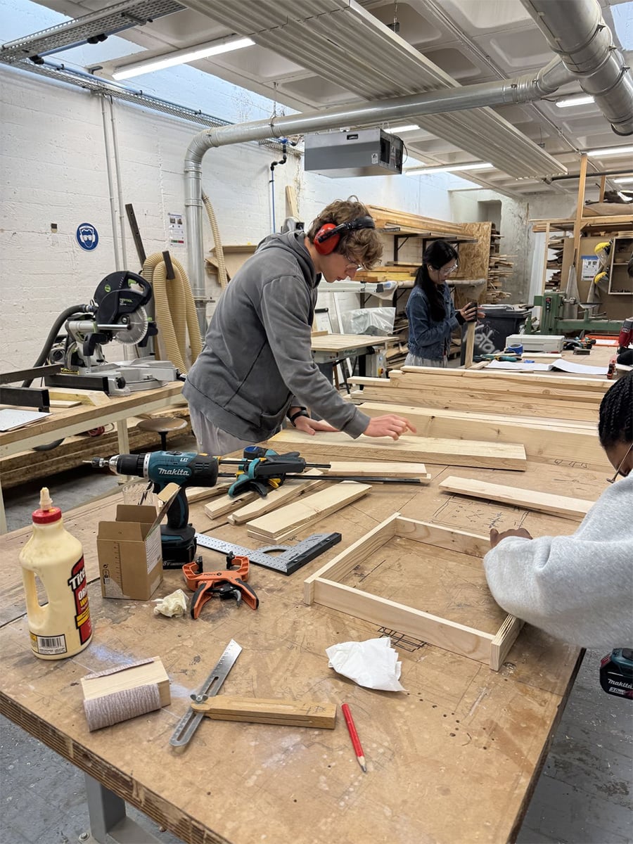 Three people work on woodworking projects in a workshop filled with wooden planks, tools, and equipment. One person wears ear protection and measures wood on a table, while others are focused on their tasks.