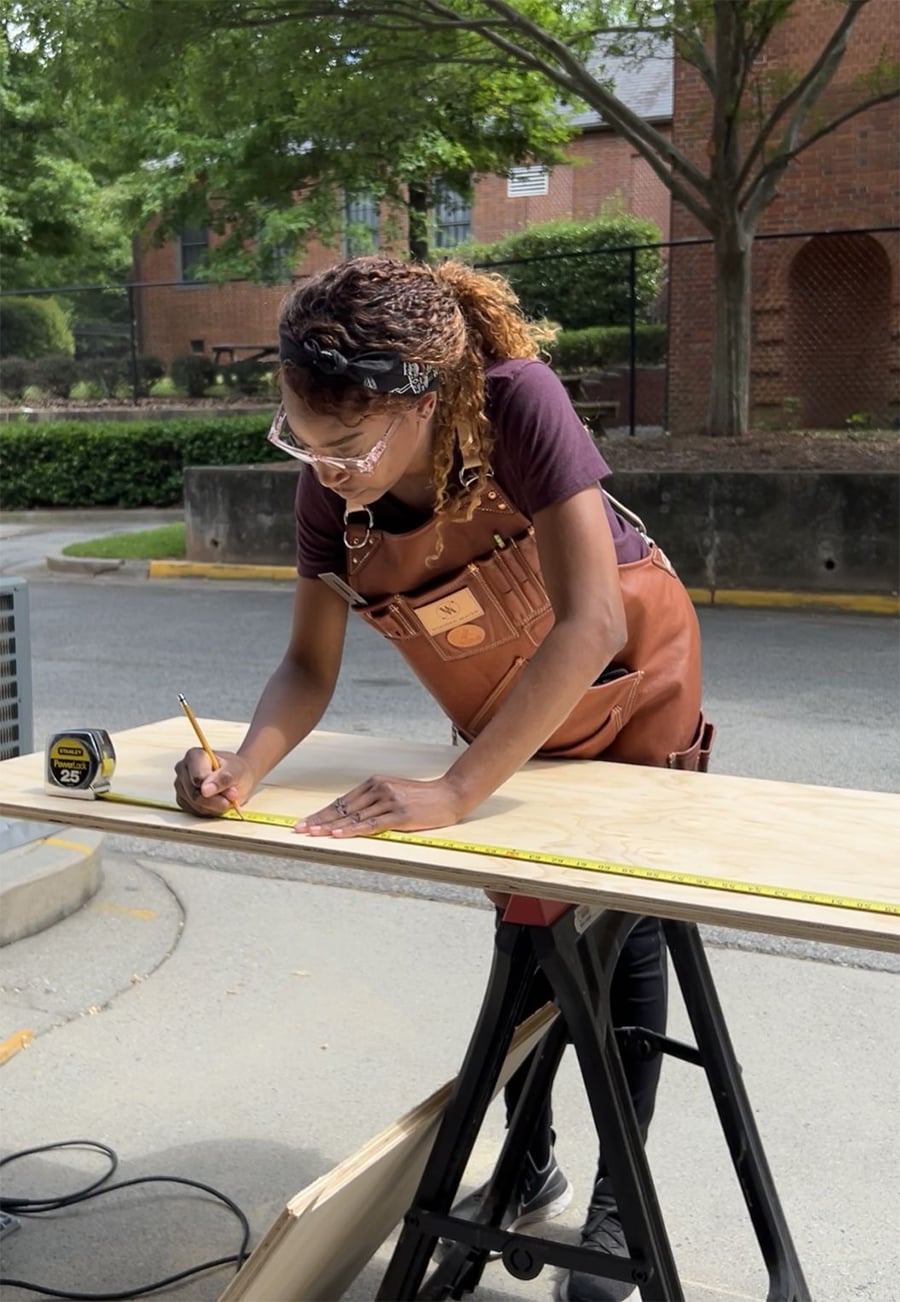 A person wearing safety glasses and a brown apron measures and marks a wooden board with a pencil and tape measure on a workbench outdoors. Trees and a brick building are in the background.