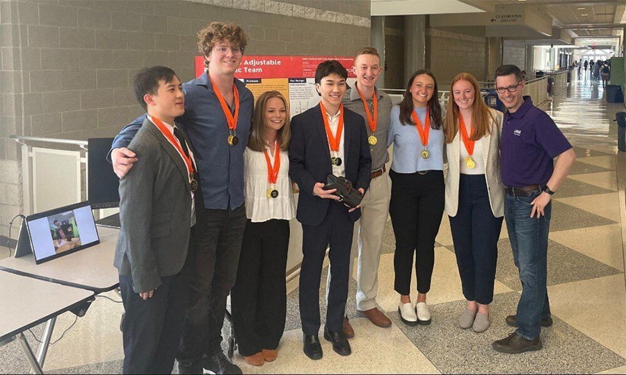 A group of seven students with medals and one adult pose together indoors, smiling. A laptop on a table nearby displays a video call. The setting appears to be a school hallway.